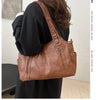 A woman holds a brown leather everyday large-capacity shoulder tote bag against a light background.