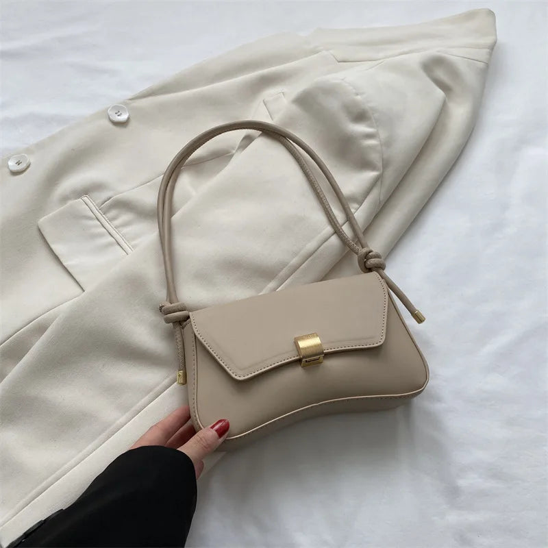A beige leather crossbody bag is shown resting on a white surface next to a cream-colored blazer.