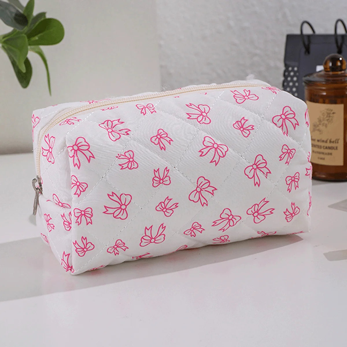 Bolsa acolchada blanca para maquillaje con estampado de lazo rosa y cremallera sobre una superficie blanca.
