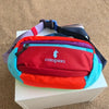 A red and blue patchwork color block fanny pack with a purple strap resting on a white box.