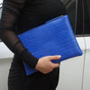A hand holds a bright blue alligator print clutch bag against a dark background.