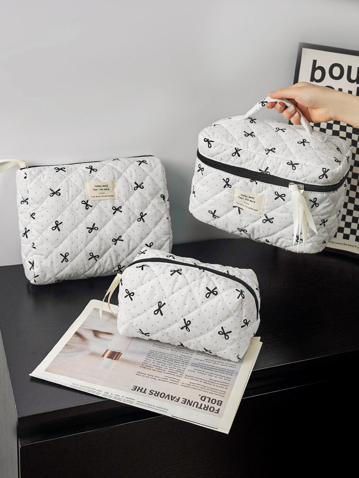 A white quilted makeup bag with black embroidery is shown next to a magazine on a dark surface.