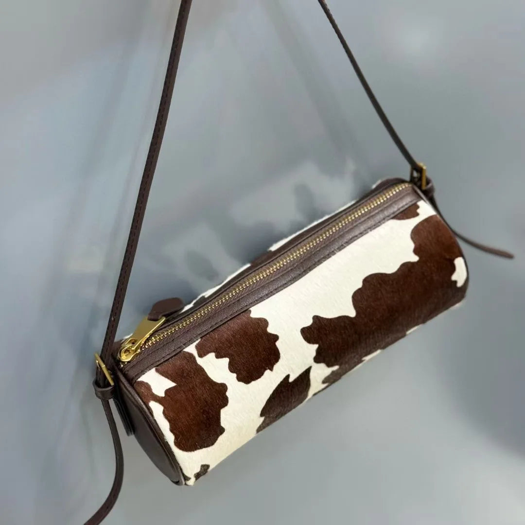 A brown and white cow print cylinder shoulder bag with a leather strap hanging against a light gray background.