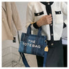 A woman holds a dark blue tote bag with white lettering against a background of a cream knit sweater and black clothing.
