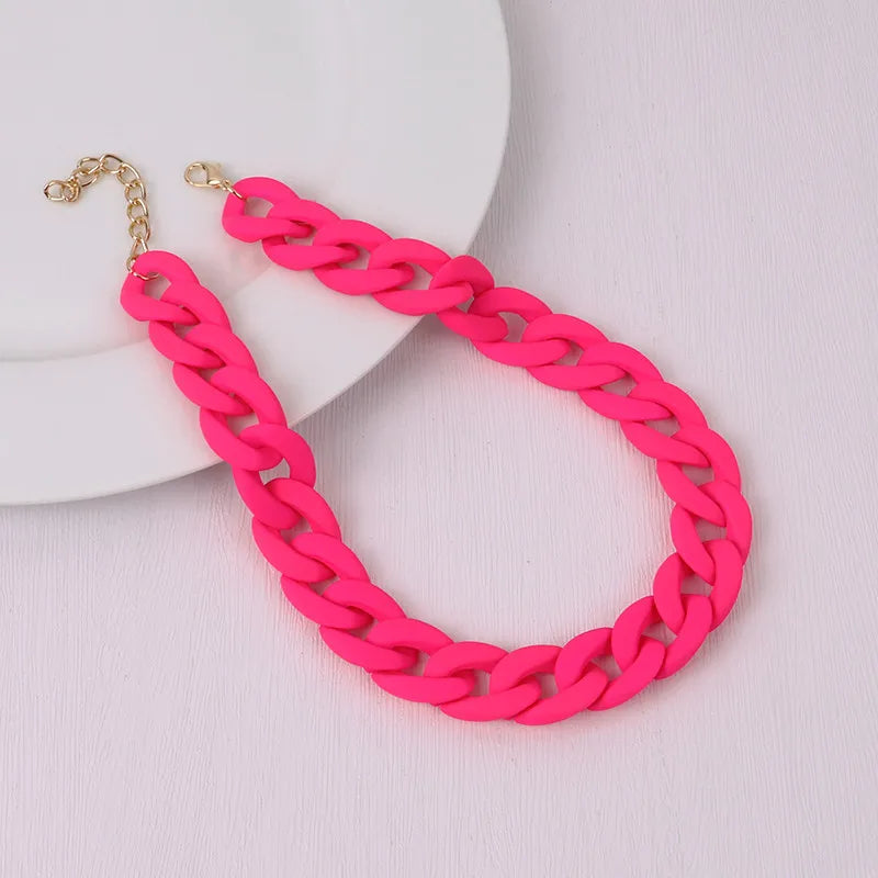 A bright pink acrylic statement choker necklace with a gold-colored chain is displayed on a white plate.