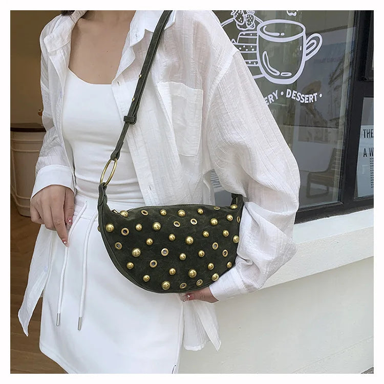 A woman wearing a white shirt and skirt holds a dark green faux suede crossbody bag with gold rivet accents.