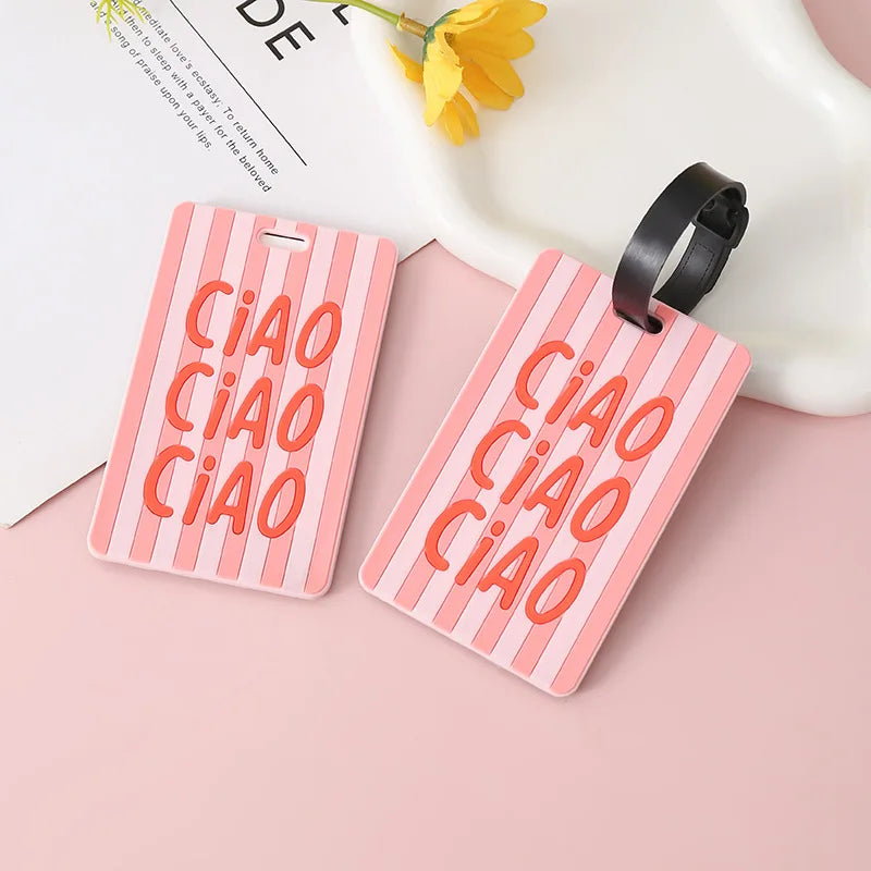 Pink silicone letter luggage tags with orange text and black strap resting on a white surface.
