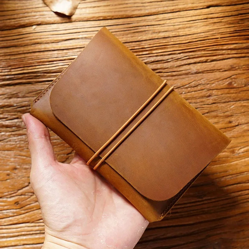 A hand holds a minimalist brown full-grain leather passport holder and card wallet on a wooden surface.