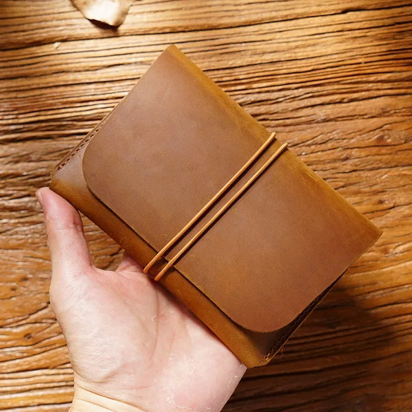 A hand holds a minimalist brown full-grain leather passport holder and card wallet on a wooden surface.