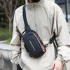 A person wearing a beige jacket is holding a black Oxford sling chest bag with a leather strap.