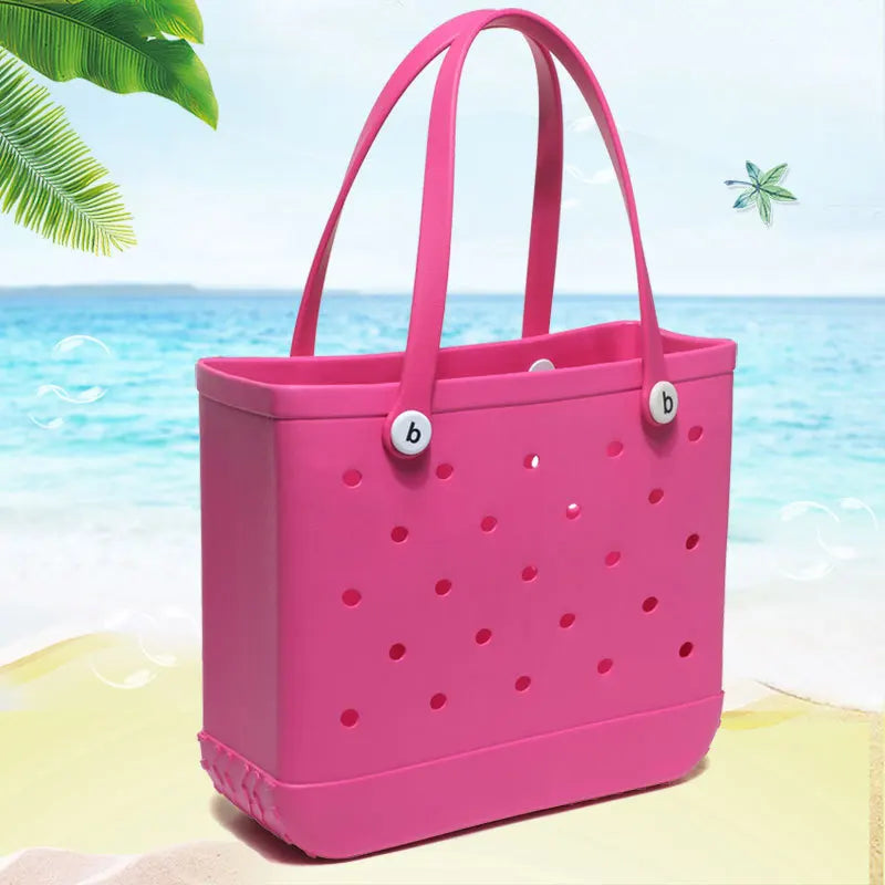 A bright pink portable EVA beach tote bag with perforated sides is shown on a sandy beach near the ocean.