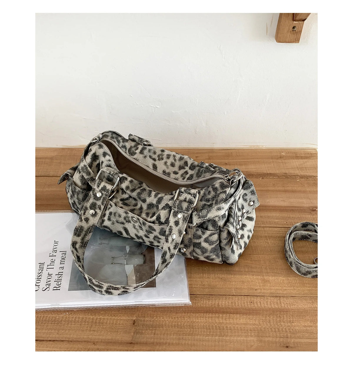 A leopard print riveted Boston shoulder bag resting on a wooden surface next to a book.