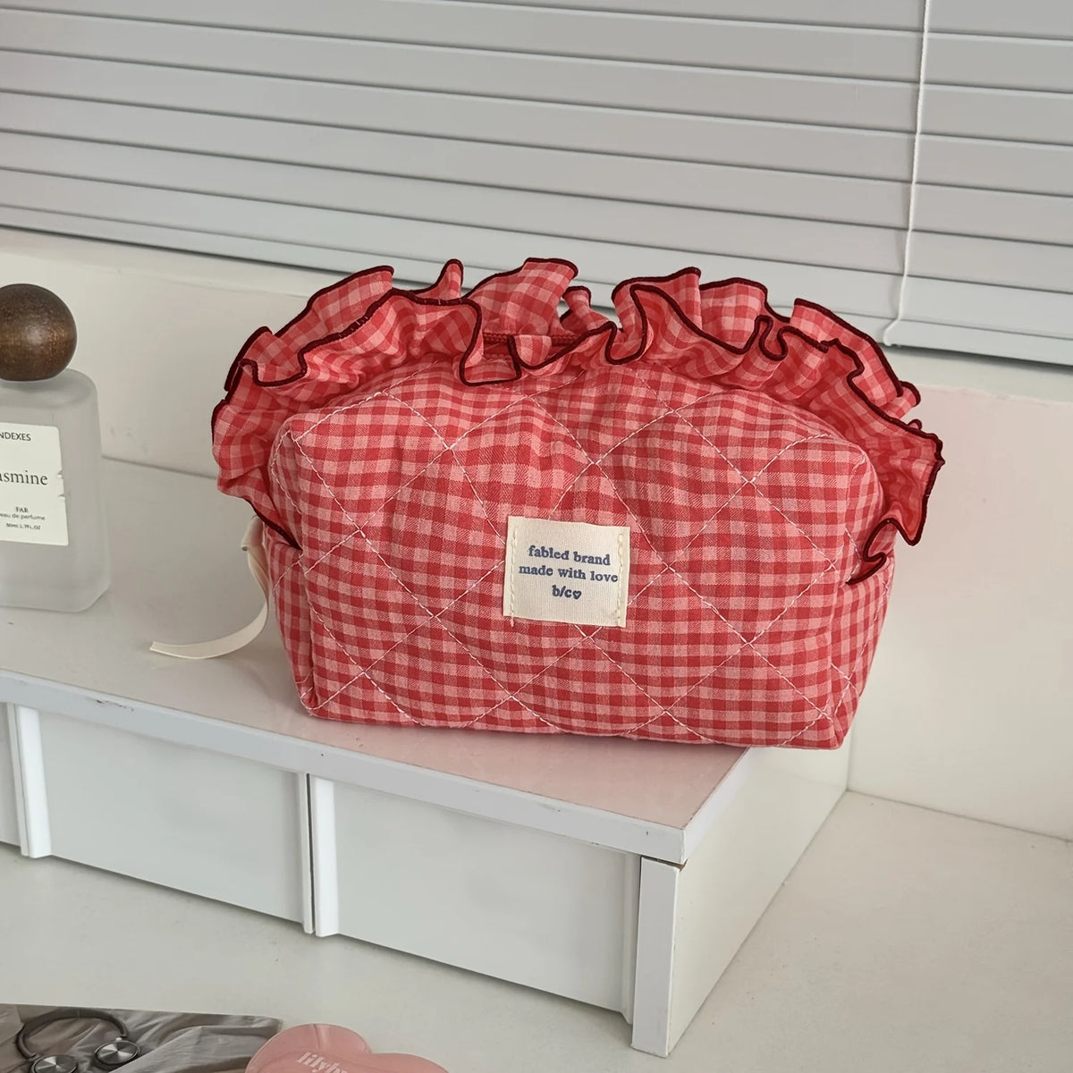 A red and white checkered makeup pouch with a ruffled lace trim resting on a white surface.