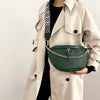 A woman wearing a beige trench coat holds a dark green woven pattern waist bag with gold chain details.