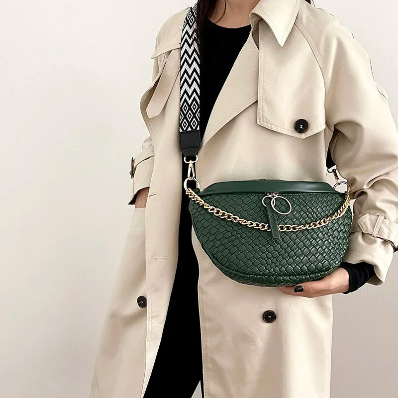 A woman wearing a beige trench coat holds a dark green woven pattern waist bag with gold chain details.