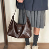 A woman wearing a dark blazer and gray skirt holds a brown leather underarm bag.