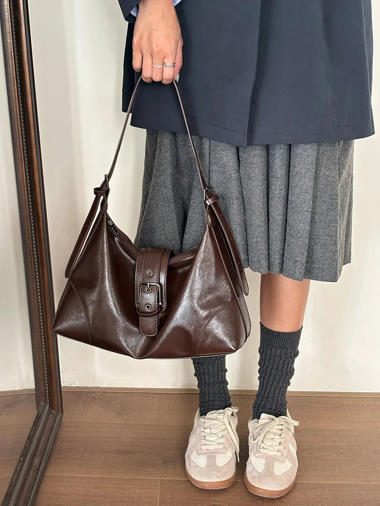 A woman wearing a dark blazer and gray skirt holds a brown leather underarm bag.