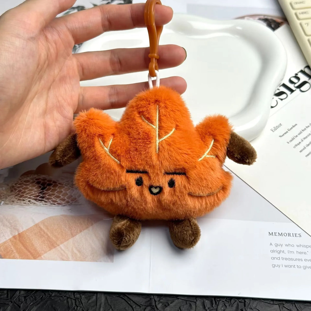 An orange maple leaf plush keychain charm is held in a hand against a white background.