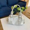 A white PU leather shoulder bag with silver rivets and a handle is displayed on a white table next to a green plant.