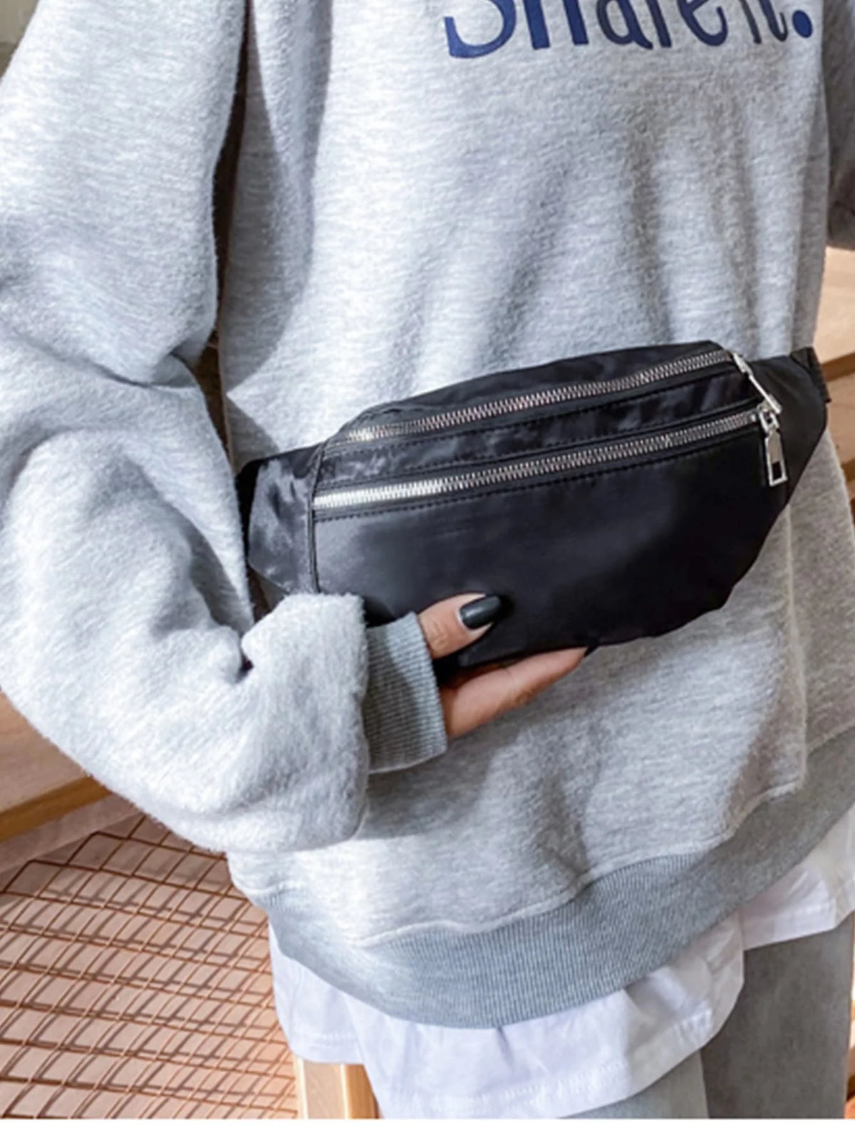 A person holds a black nylon crossbody sling bag against a gray sweatshirt.