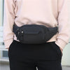 A black canvas waist fanny bag is shown being held by a person wearing a light pink top and black pants.