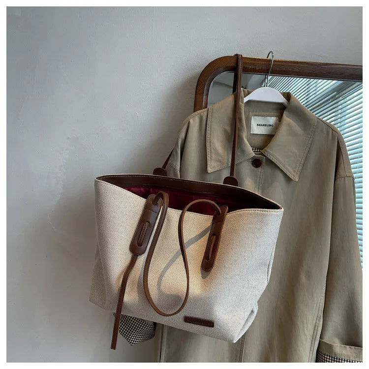 A beige canvas shoulder tote bag is displayed hanging on a wooden hanger against a white wall.