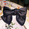 A black silk bow hair clip is displayed on a white surface with green leaves.