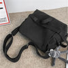 Bolso cruz de lona negra con un peluche gris adjunto, sobre una superficie beige.