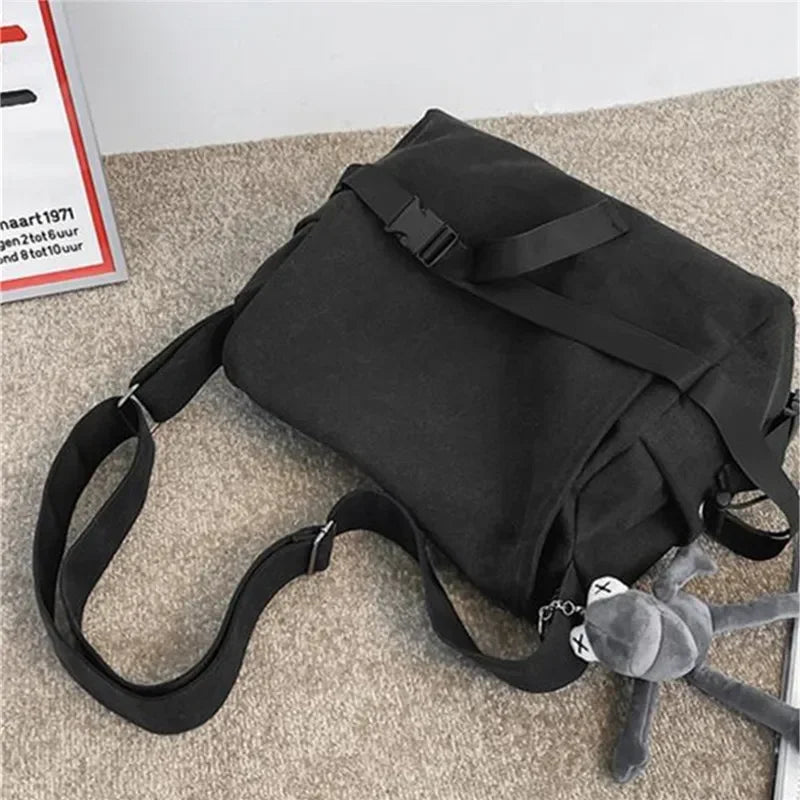 Bolso cruz de lona negra con un peluche gris adjunto, sobre una superficie beige.