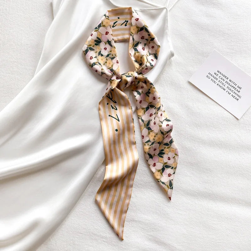Foulard pour cheveux en soie, motif floral jaune et rose, noué en nœud, posé sur tissu blanc.