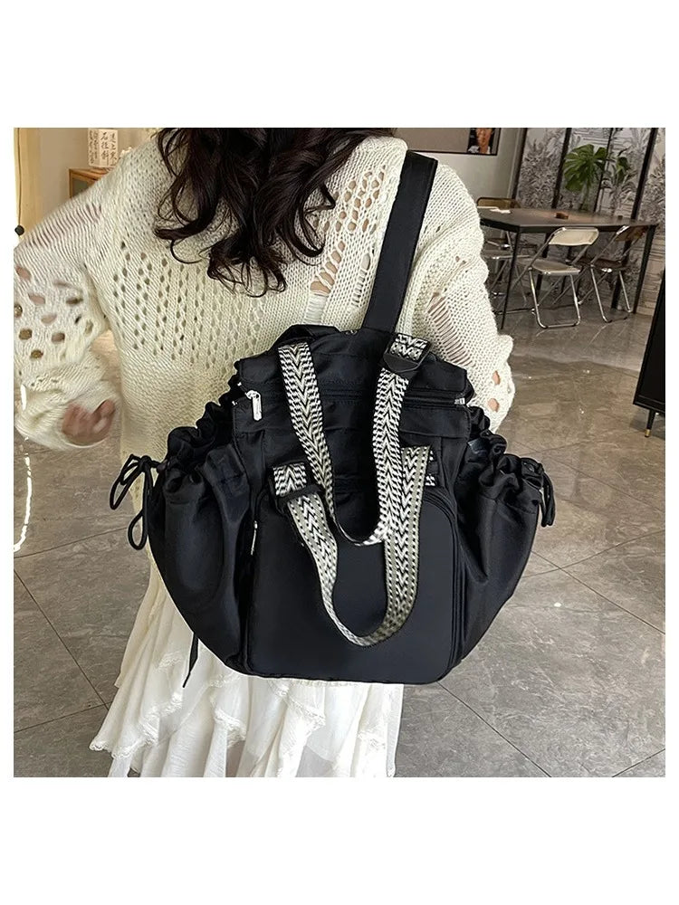 A black nylon shoulder bag with a silver chain strap is carried by a woman wearing a cream knit sweater and white skirt.