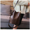 A brown alligator pattern leather crossbody bag is displayed hanging from a person's hand in front of a gray sofa.