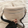 A beige soft leather crossbody bag with double zippers and a black adjustable strap resting on white bedding.