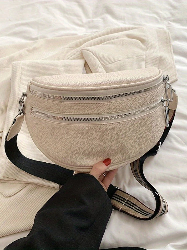 A beige soft leather crossbody bag with double zippers and a black adjustable strap resting on white bedding.