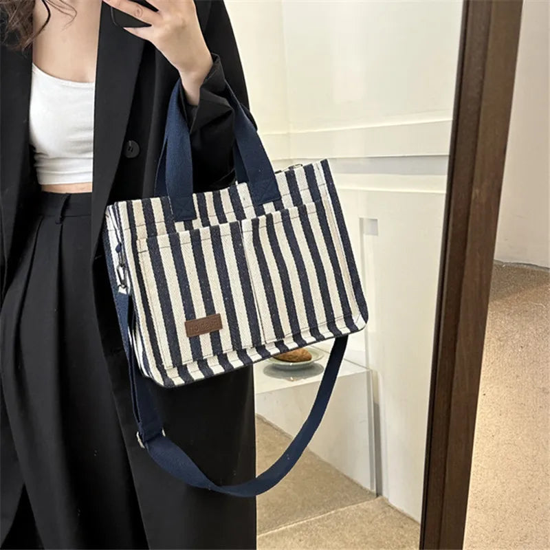 Une femme tient un sac bandoulière à toile rayé marine et blanc contre un manteau noir.