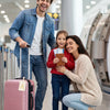 Une famille de trois souriants debout dans un aéroport avec une valise à roulettes.