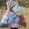 A person holds a large, colorful canvas shopping tote featuring an impressionistic cityscape painting.