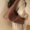 A woman is holding a brown suede shoulder tote bag against a light-colored shirt.