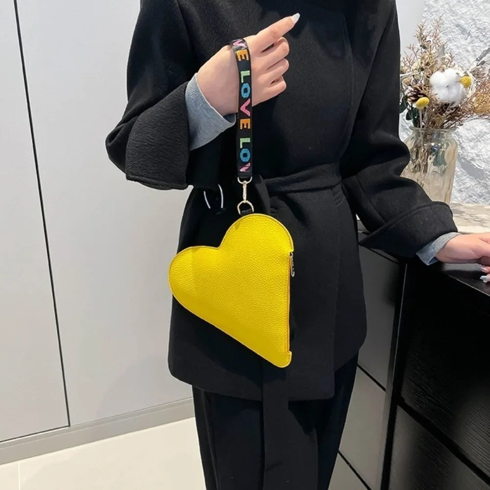 Cartera de monedas de forma de corazón amarilla y cuero PU con correa negra, sostenida por una persona con abrigo negro.