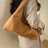 A woman is holding a tan suede shoulder tote bag over her shoulder, featuring a relaxed, neutral outfit.