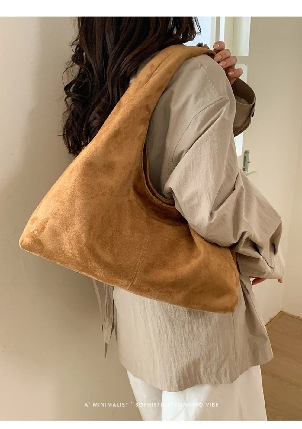 A woman is holding a tan suede shoulder tote bag over her shoulder, featuring a relaxed, neutral outfit.