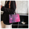 A woman holds a pink and purple patterned ladies' shoulder tote bag with a brown strap against a dark background.