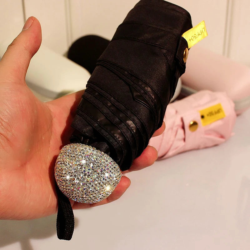 A hand holds a black compact umbrella with a rhinestone sphere and pink fabric, labeled for UV protection.