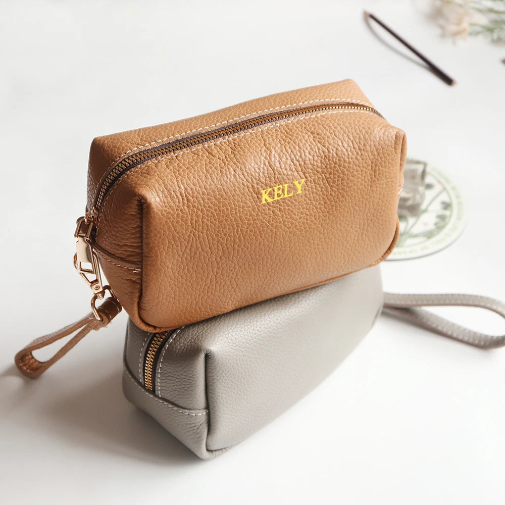 A tan leather card holder and coin purse with gold stitching resting on a gray leather pouch.