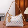 A beige leather crescent tote bag with a white and pink letter print and a brown bow accent resting on a wooden surface.