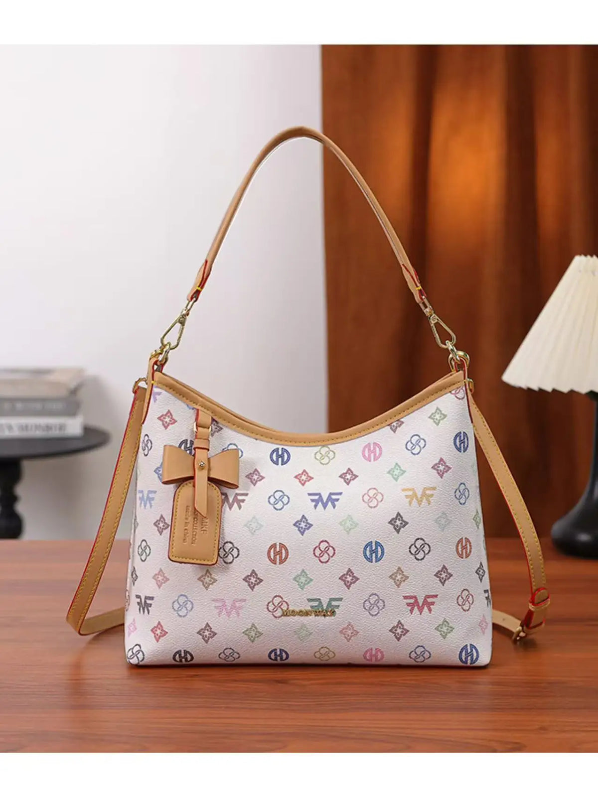 A beige leather crescent tote bag with a white and pink letter print and a brown bow accent resting on a wooden surface.