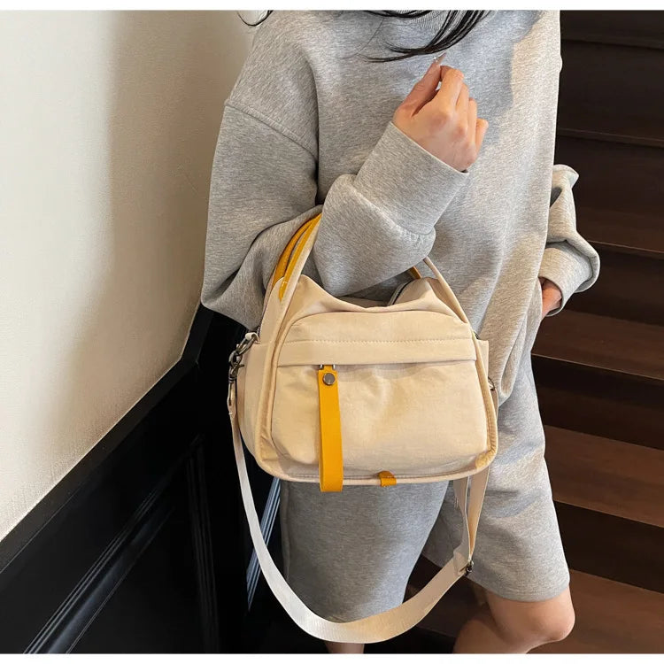 A light beige nylon crossbody bag with a top handle is shown being held by a person in a gray sweater.