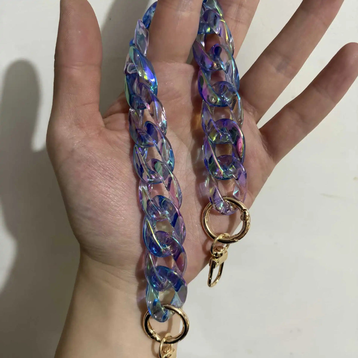 A hand holds a long, iridescent blue resin chain strap with a gold clasp.