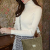Une femme portant un pull col roulé blanc et une jupe à motifs tient un sac à épaule en velours côtelé vert.