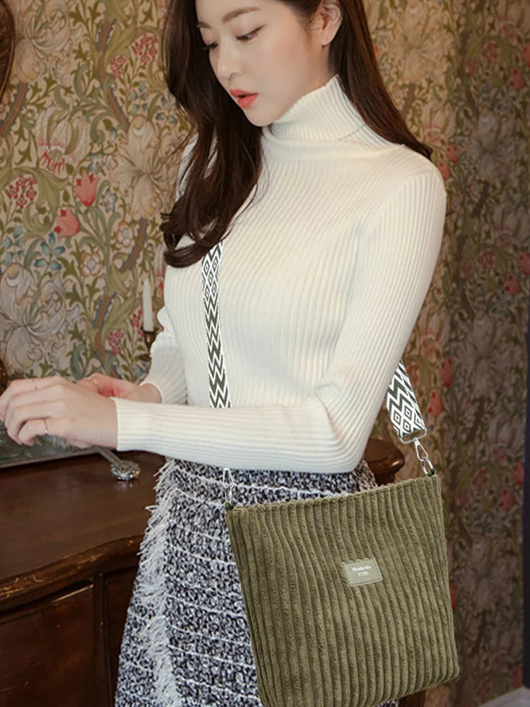 Une femme portant un pull col roulé blanc et une jupe à motifs tient un sac à épaule en velours côtelé vert.